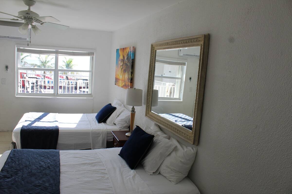 A hotel room with two beds, a ceiling fan, a large window with a view of palm trees, and a painting on the wall.