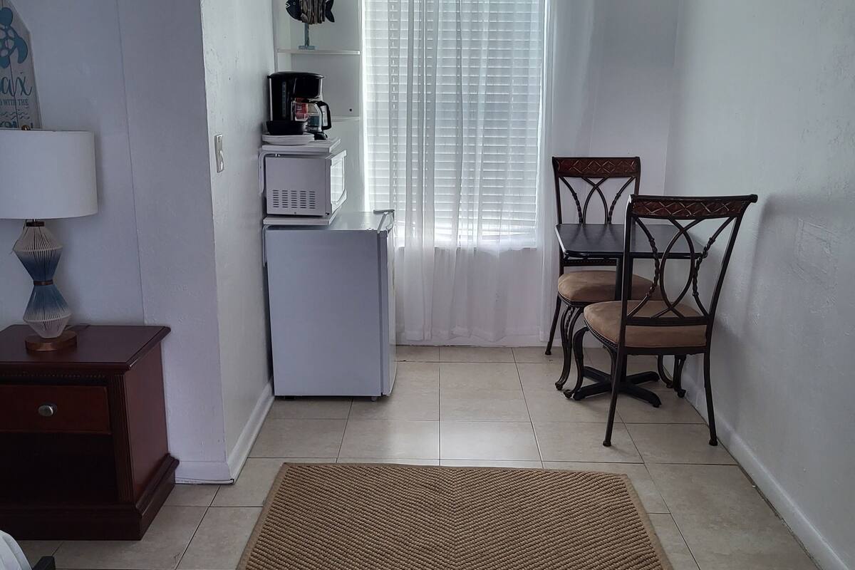 A small, clean room with a kitchenette, a dining area with two chairs, a rug, and a bed.