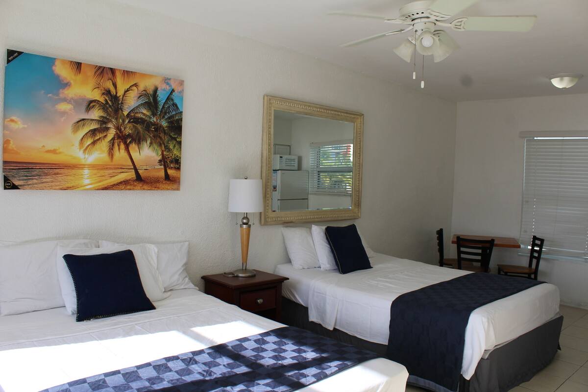 A hotel room with two beds, a ceiling fan, a painting of a sunset, a mirror, and a window with blinds.