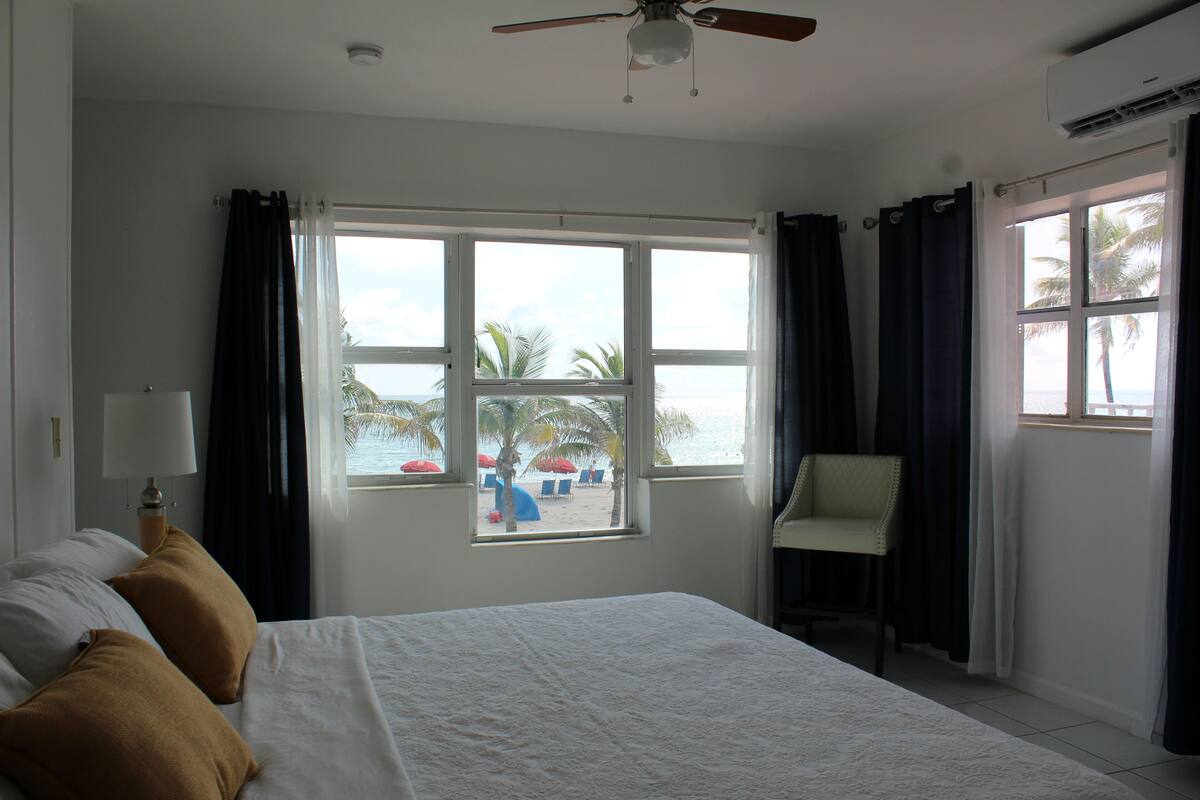 A hotel room with a bed, a chair, and a window with a view of the beach and palm trees.
