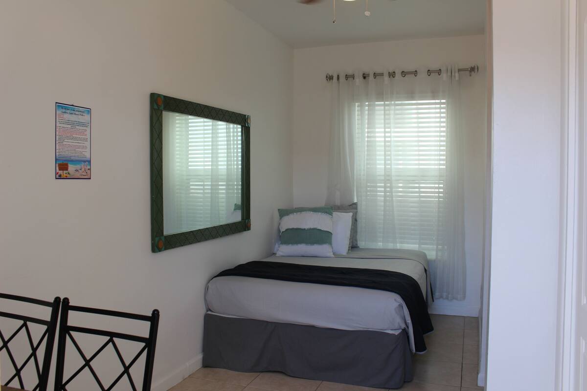 A bedroom with a bed, a mirror, a window with blinds, and a door.