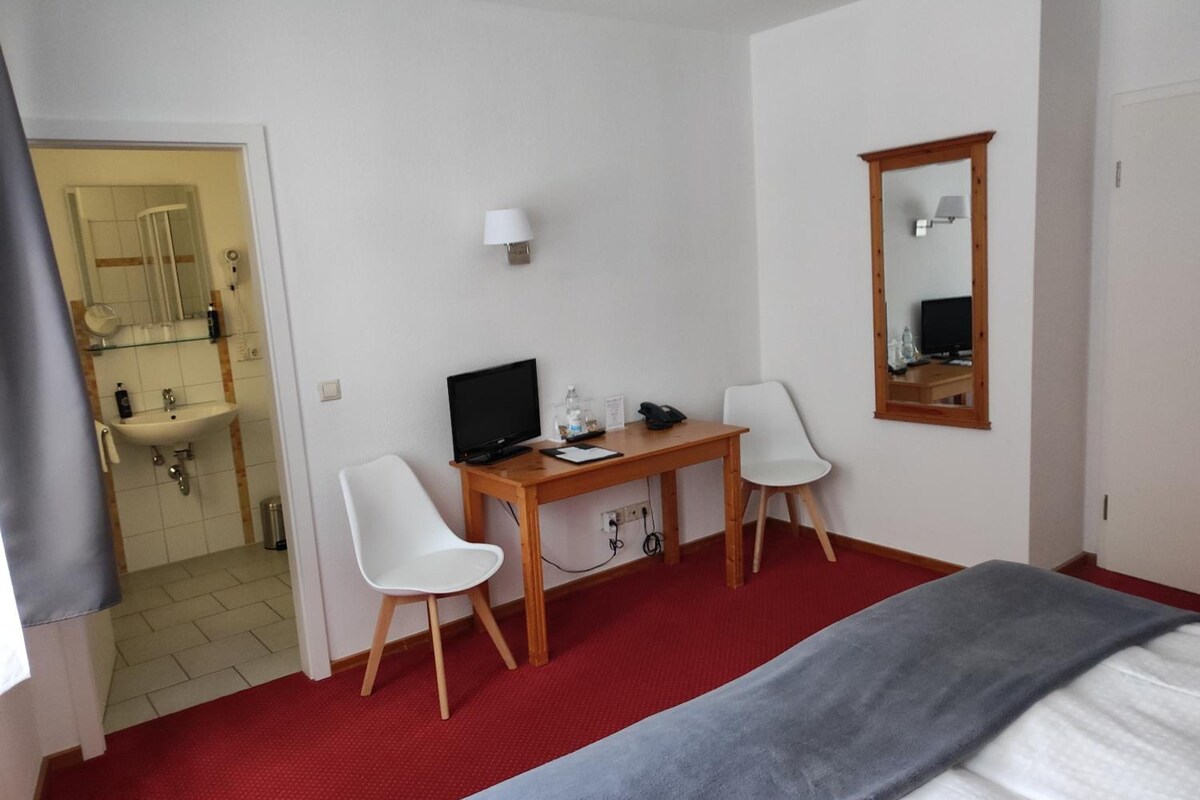 A hotel room with a bed, a desk with a computer, two chairs, a mirror, and a bathroom visible through an open door.