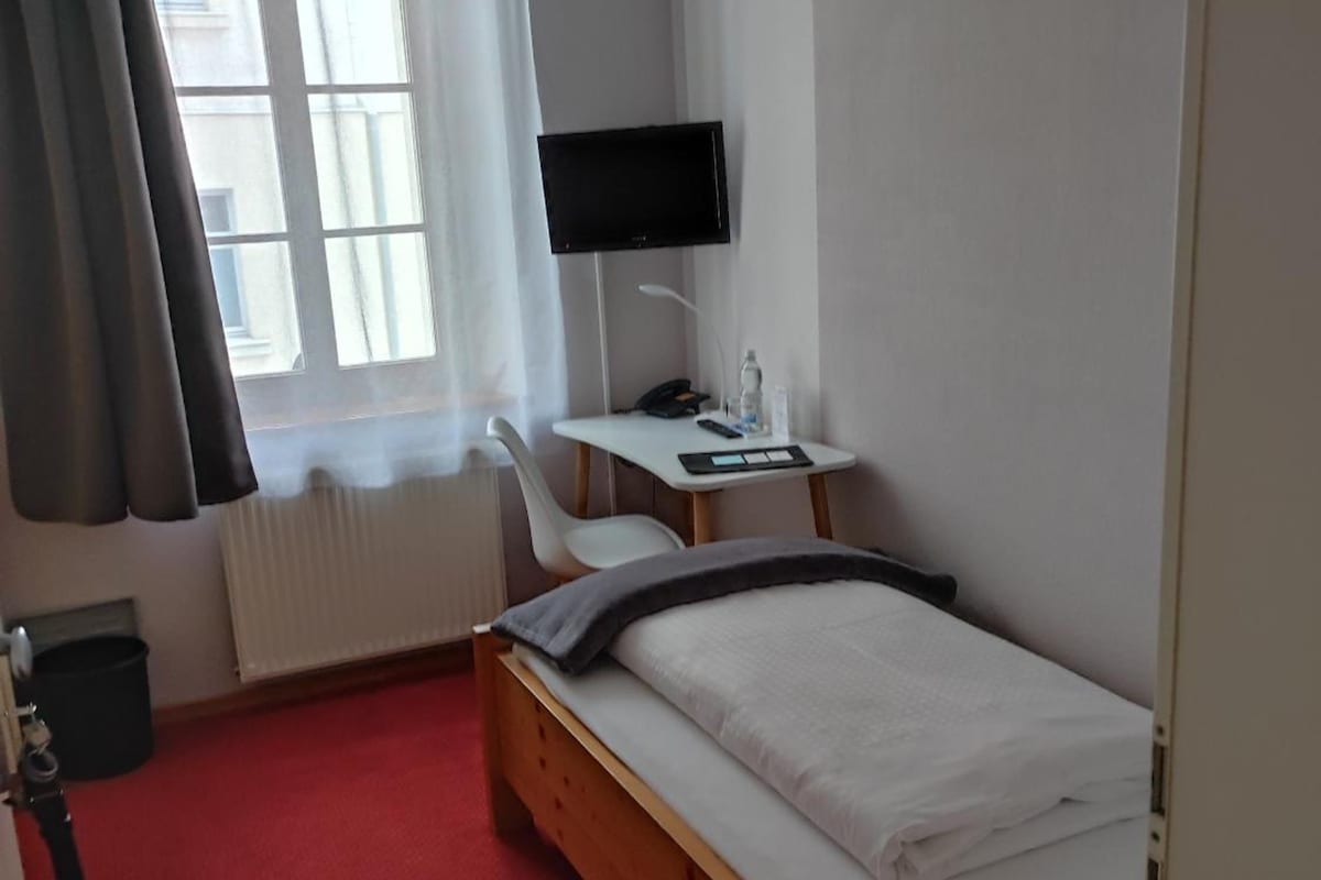 Habitación de hotel con una cama individual, un escritorio con computadora, una silla, un televisor montado en la pared y una ventana con cortinas.