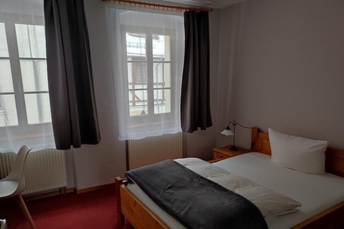 Habitación de hotel con una cama, una silla, una mesita de noche con lámpara y una ventana con cortinas.