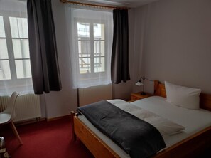 A hotel room with a bed, a chair, a nightstand with a lamp, and a window with curtains.