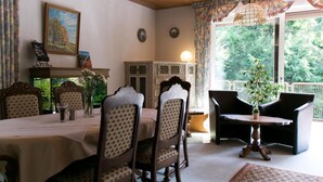 A dining area with a table set for four, surrounded by patterned chairs. There is a cabinet with glass doors, a painting on the wall, and a window with curtains. - Haus am Wasserfall (Detmold)