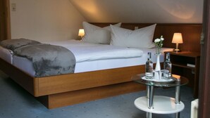 A modern hotel room with a large bed, two bedside lamps, a small round table with a vase and bottles, and a wooden headboard. - Haus am Wasserfall (Detmold)