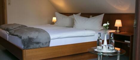 Una habitación de hotel moderna con una cama grande, dos lámparas de noche, una mesita redonda con un florero y botellas, y un cabecero de madera.