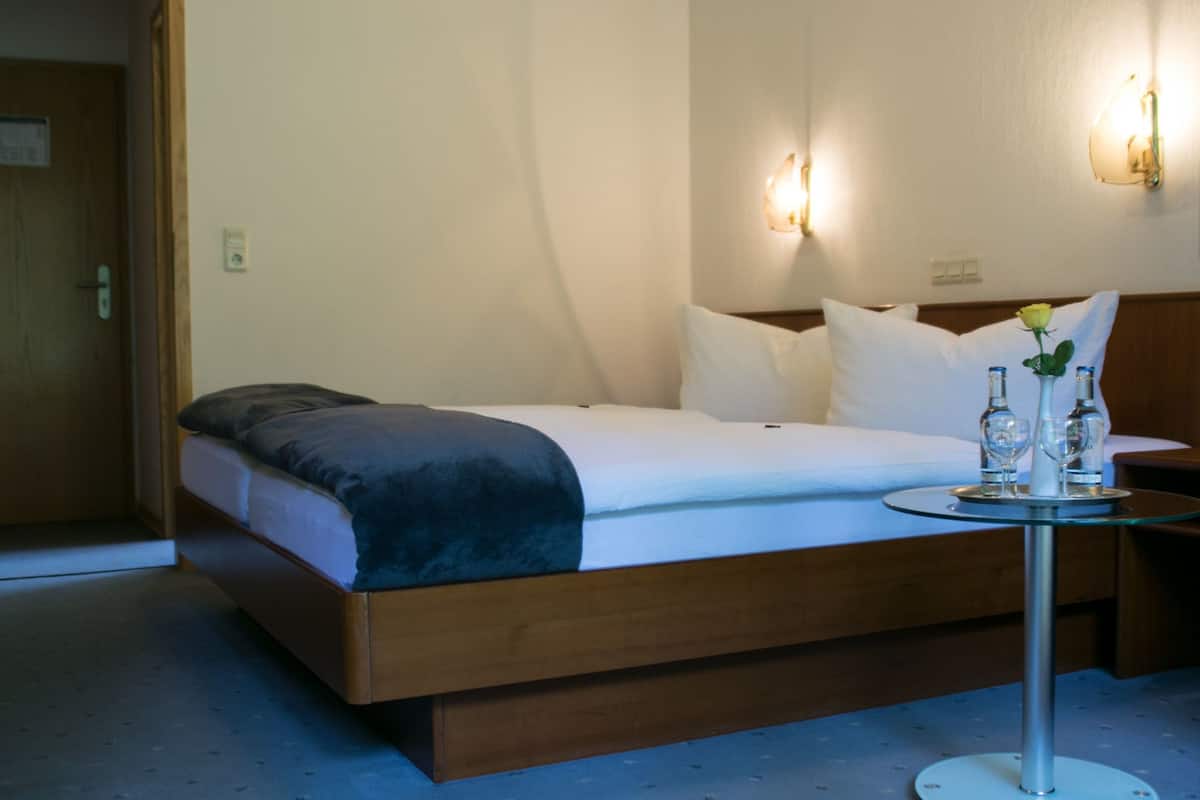 Habitación de hotel con una cama individual, una mesita de noche con un florero y una pequeña mesa con una botella y un vaso.