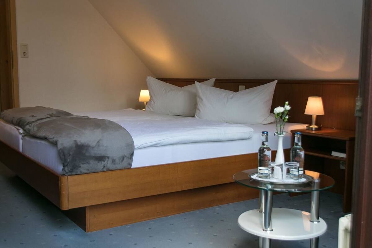 Habitación de hotel con una cama grande, una mesita de noche con lámpara y una pequeña mesa redonda con botellas y un florero.