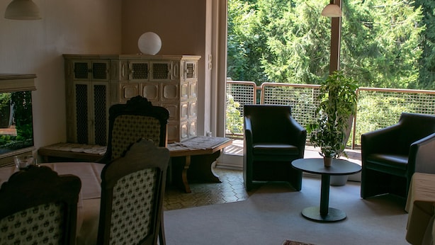 A cozy living room with a fireplace, a large window with a view of trees, and a balcony with chairs and a table. - Haus am Wasserfall (Detmold)