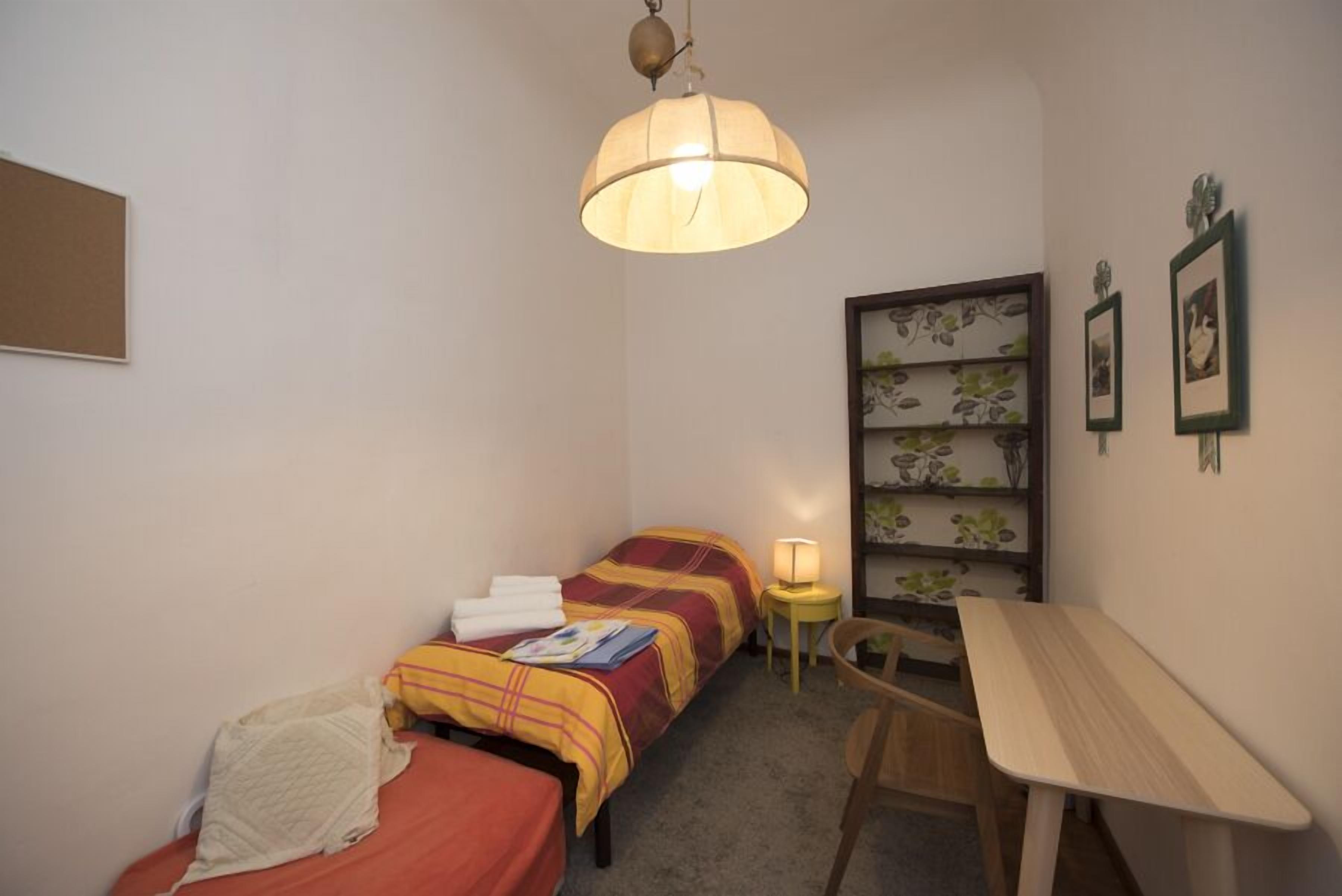 Une petite pièce agréable, dotée d’un lit simple, d’un bureau, d’une chaise, d’une bibliothèque et d’une lampe suspendue.