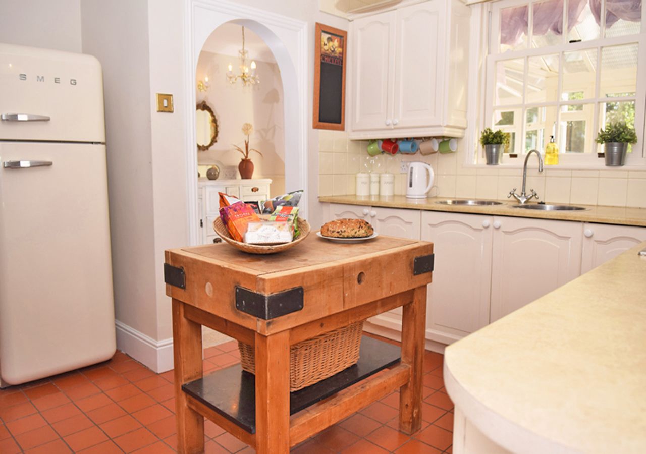 A kitchen with white cabinets, a white refrigerator, a wooden table with a basket, and a window with plants.