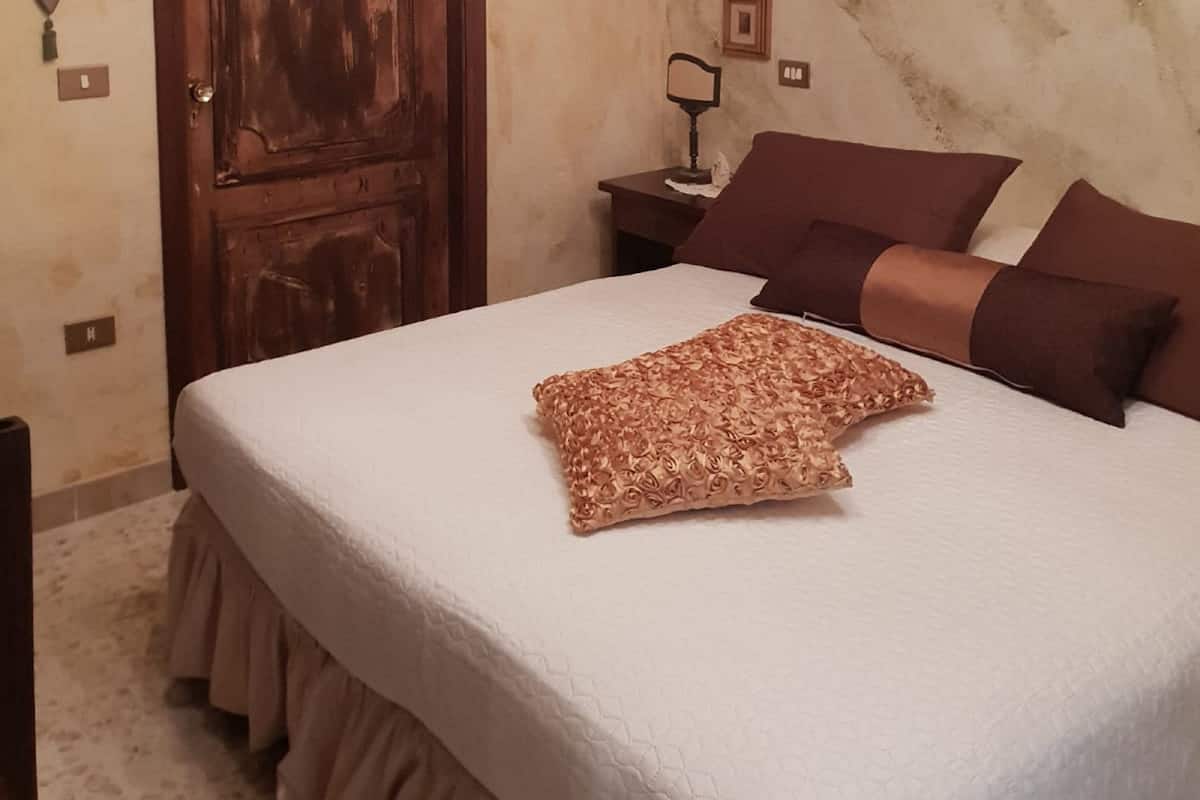 Une chambre avec une porte en bois, un lit agrémenté d’un coussin décoratif, une table de chevet avec une lampe et un tableau accroché au mur.