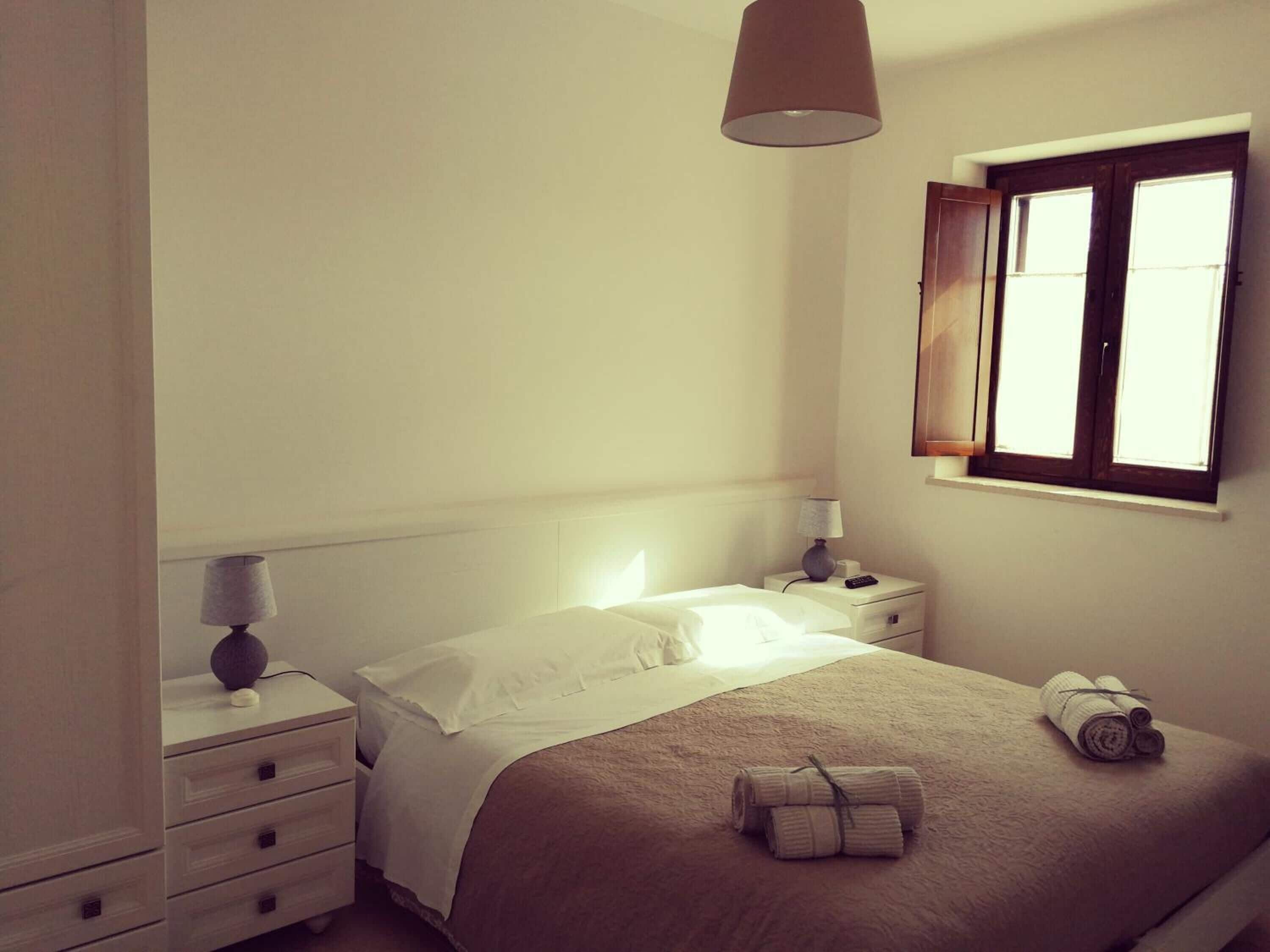 Una cama bien hecha con edredón blanco y manta marrón, una lámpara de noche, una mesita con un control remoto y una ventana con cortinas.