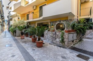Una calle urbana estrecha con macetas a lo largo de la acera, un muro de piedra con un ventanal circular y un edificio amarillo con balcones.