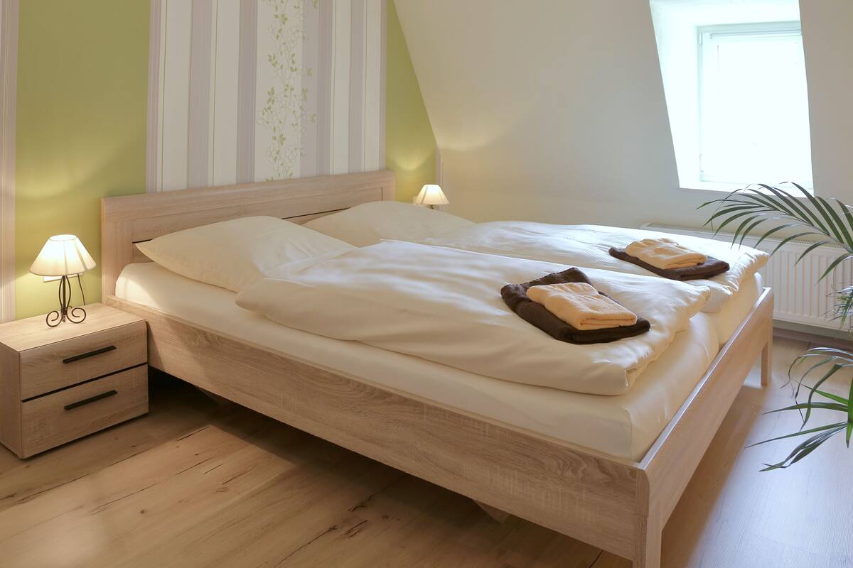 A neatly made bed with two pillows and two folded towels, a bedside table with a lamp, and a potted plant beside the window.