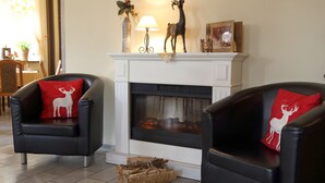 A living room with two black leather armchairs, each with a red pillow featuring a white deer design. A white fireplace with a decorative deer figurine on top is in the center. - Pension Gasthof und Hotel Roter Hirsch (Claussnitz)