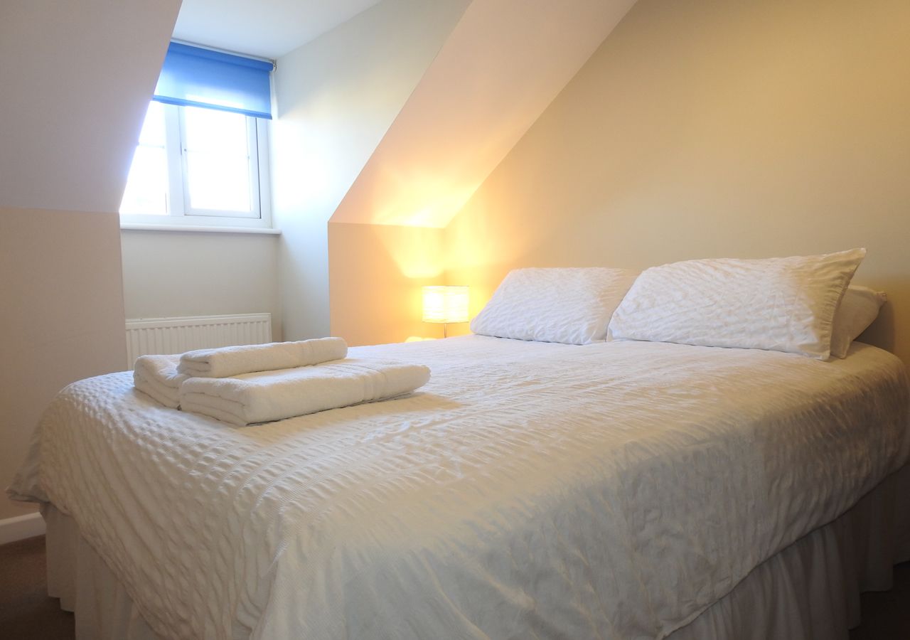 Een net opgemaakt bed met witte lakens en kussens, een bedlampje en een raam met een blauwe blind.