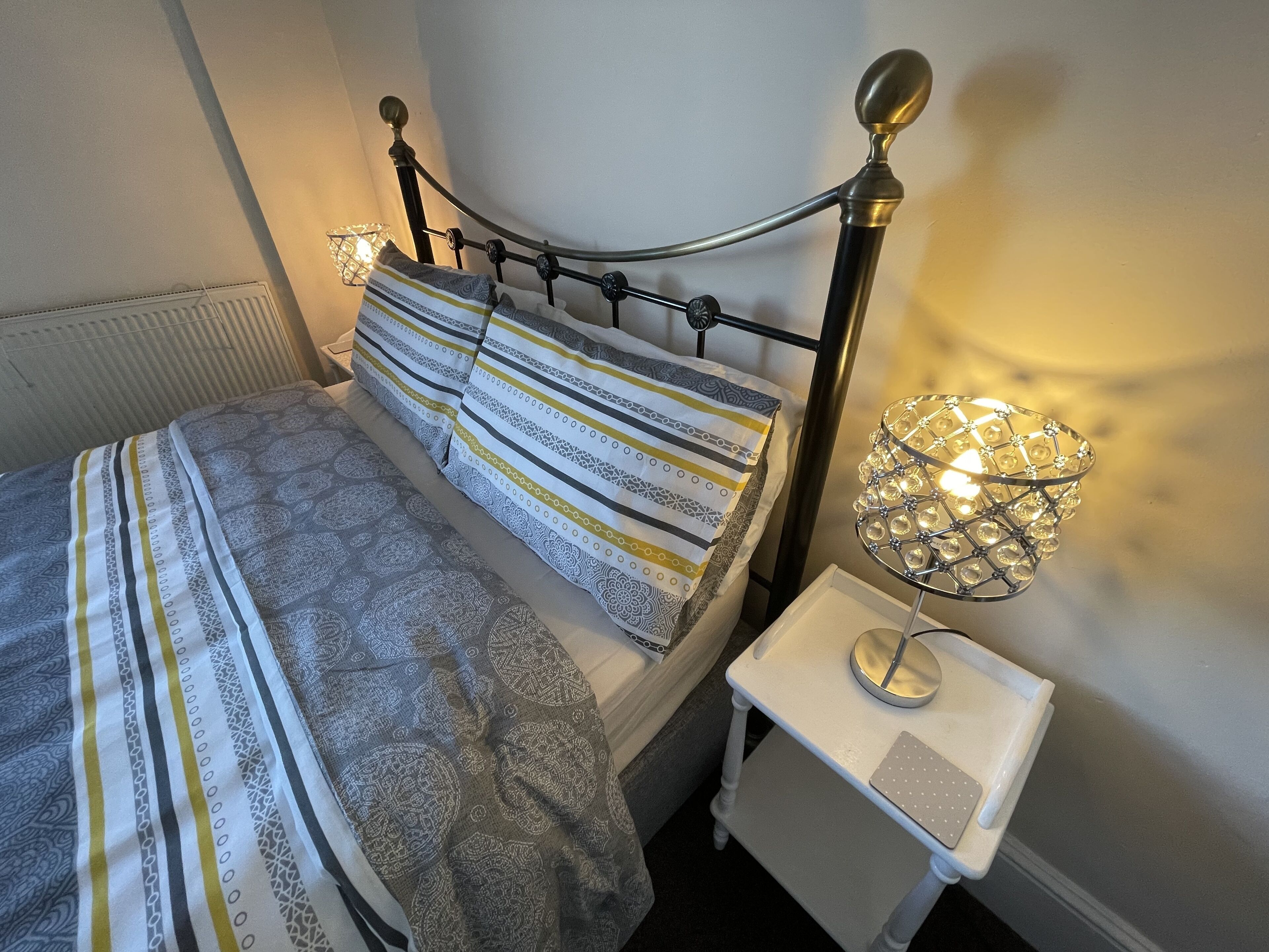 Una cama bien hecha con ropa de cama estampada, una mesita de noche con lámpara y un radiador al fondo.