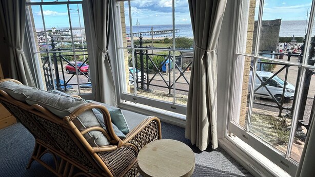 A room with a wicker chair and a small table, large windows with curtains, and a view of a coastal area with cars and a pier. - Bon-ami (Lowestoft)