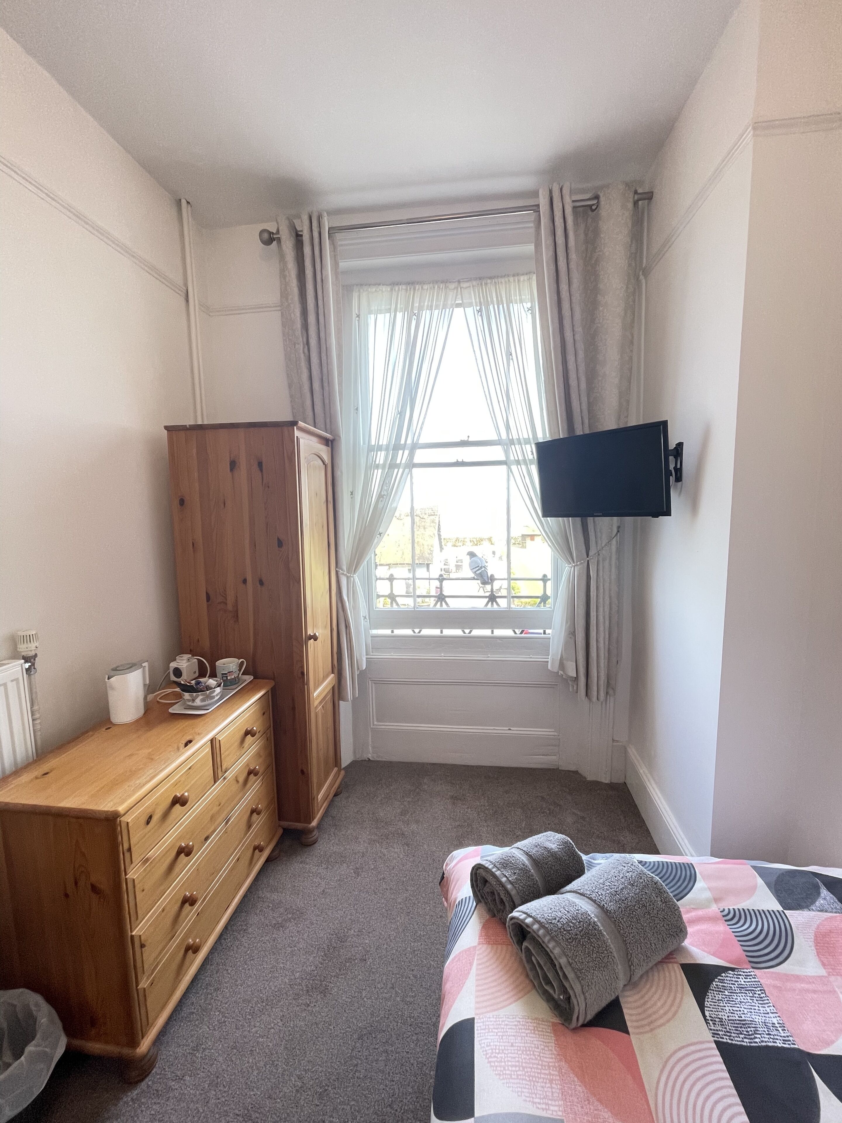 Un dormitorio con un aparador de madera, una cama con cubrecama estampado, una ventana con cortinas y un televisor de pared.