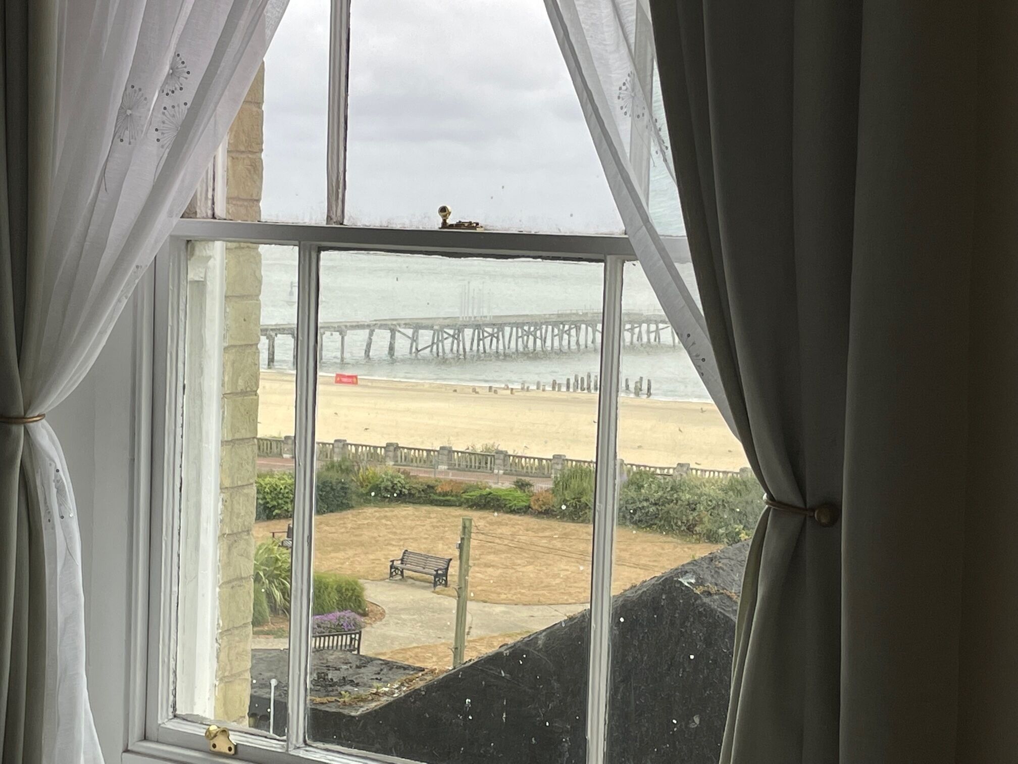 A view through a window with curtains, overlooking a beach and a pier.