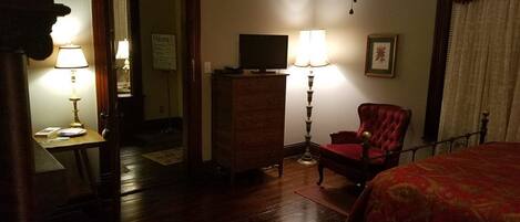 Une chambre d’hôtel avec un plancher en bois, un lit recouvert d’une couverture rouge, un fauteuil rouge, une commode, un téléviseur posé sur la commode et deux lampes de chaque côté de la commode.