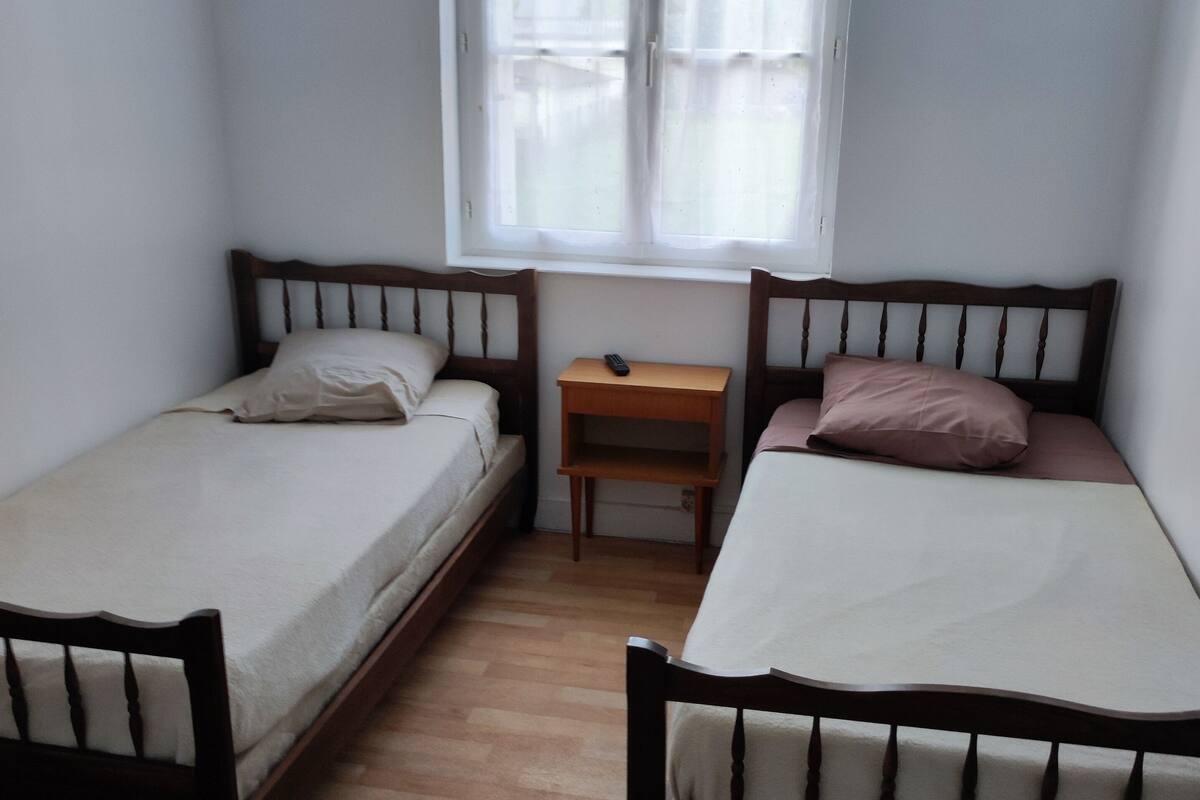 A small, clean room with two single beds, a wooden nightstand, and a window with sheer curtains.