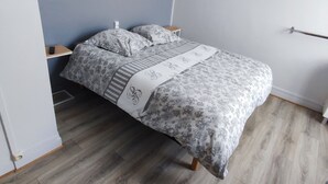 A single bed with a floral-patterned bedspread, a bedside table with a lamp, and a wooden floor. - Hôtel Au Bon Coin (Colleville)