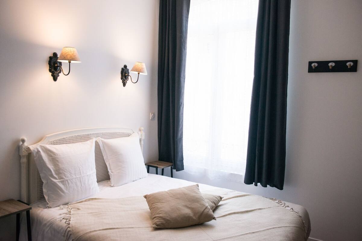 Cama bem arrumada com lençóis brancos e travesseiro bege, criado-mudo de madeira pequeno e duas luminárias fixadas na parede.