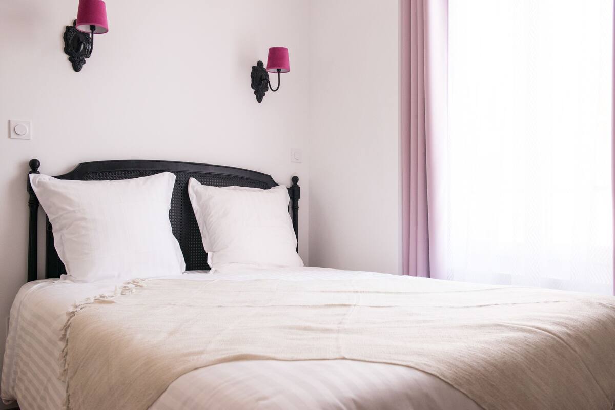 Cama bem arrumada com roupa de cama branca e cabeceira preta. Há duas luminárias fixadas na parede com cúpulas cor-de-rosa.