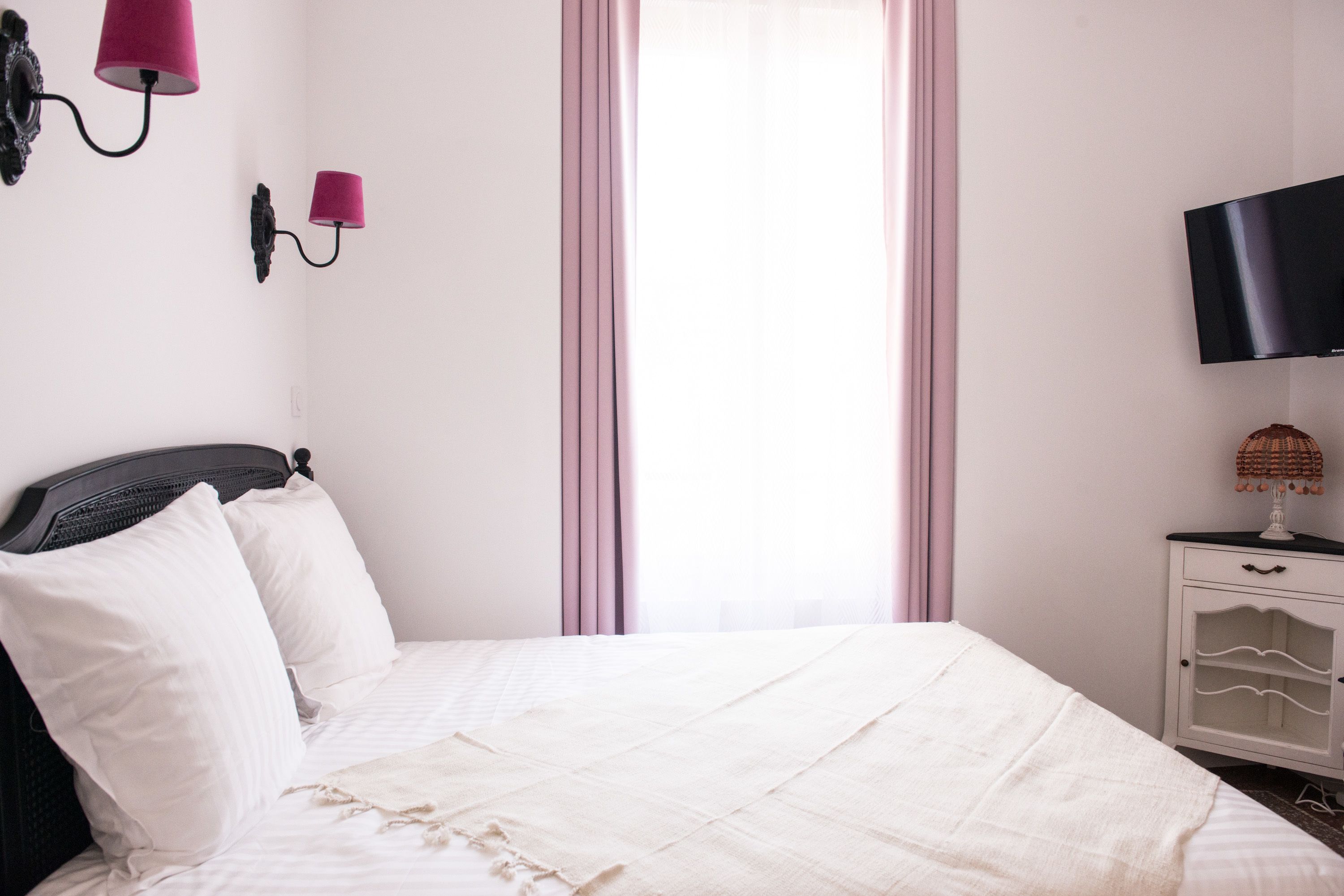 Un lit bien fait, avec du linge de lit blanc et une tête de lit, une table de chevet avec une lampe et un téléviseur fixé au mur.