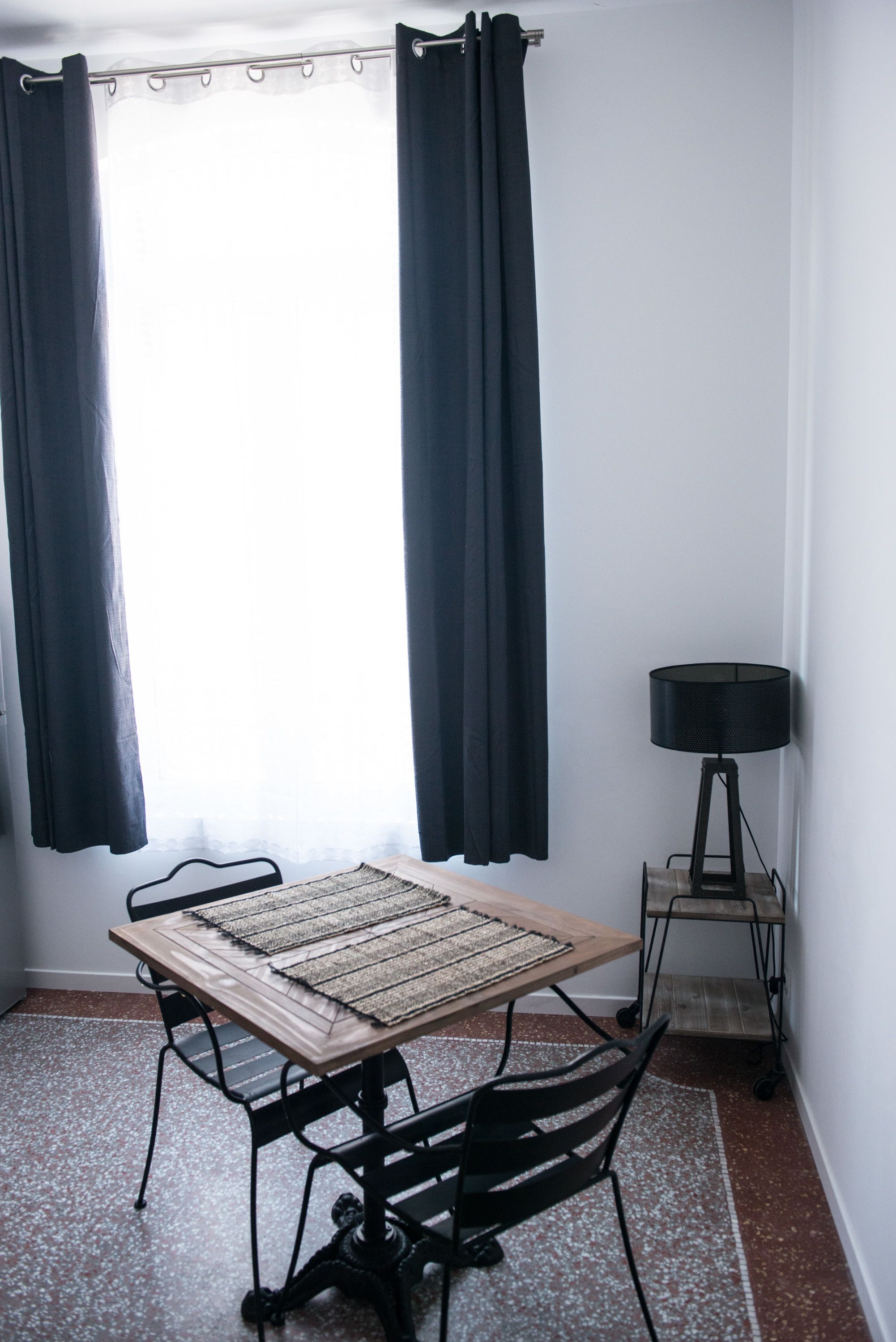 Et lille rum med et bord, stole, en lampe og et vindue med gardiner.