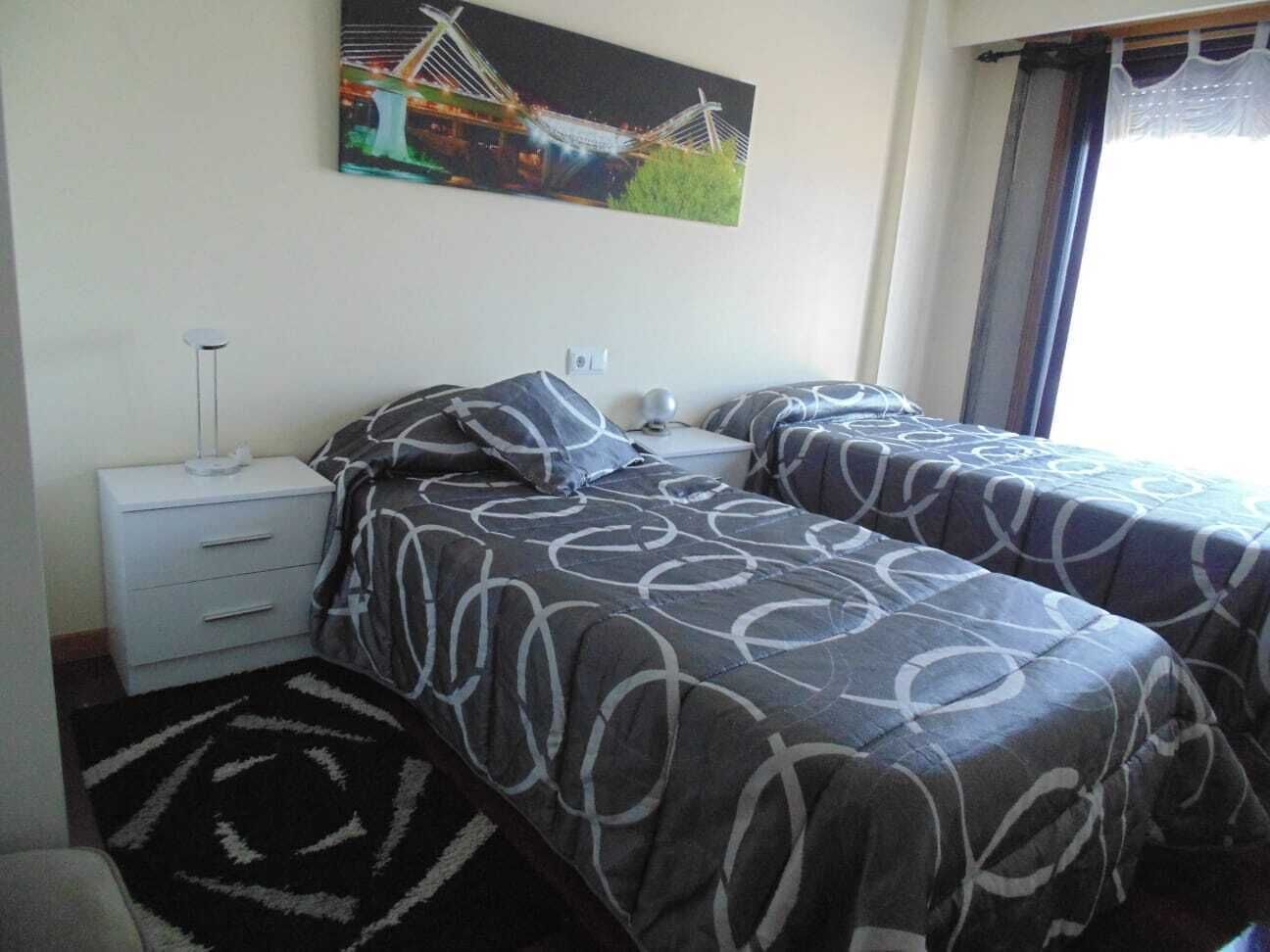 A hotel room with two beds, a nightstand, a lamp, and a painting of a bridge.