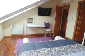 Une chambre avec un plafond en pente, un lit avec une literie à motifs, un bureau avec une chaise, un téléviseur fixé au mur et une porte.