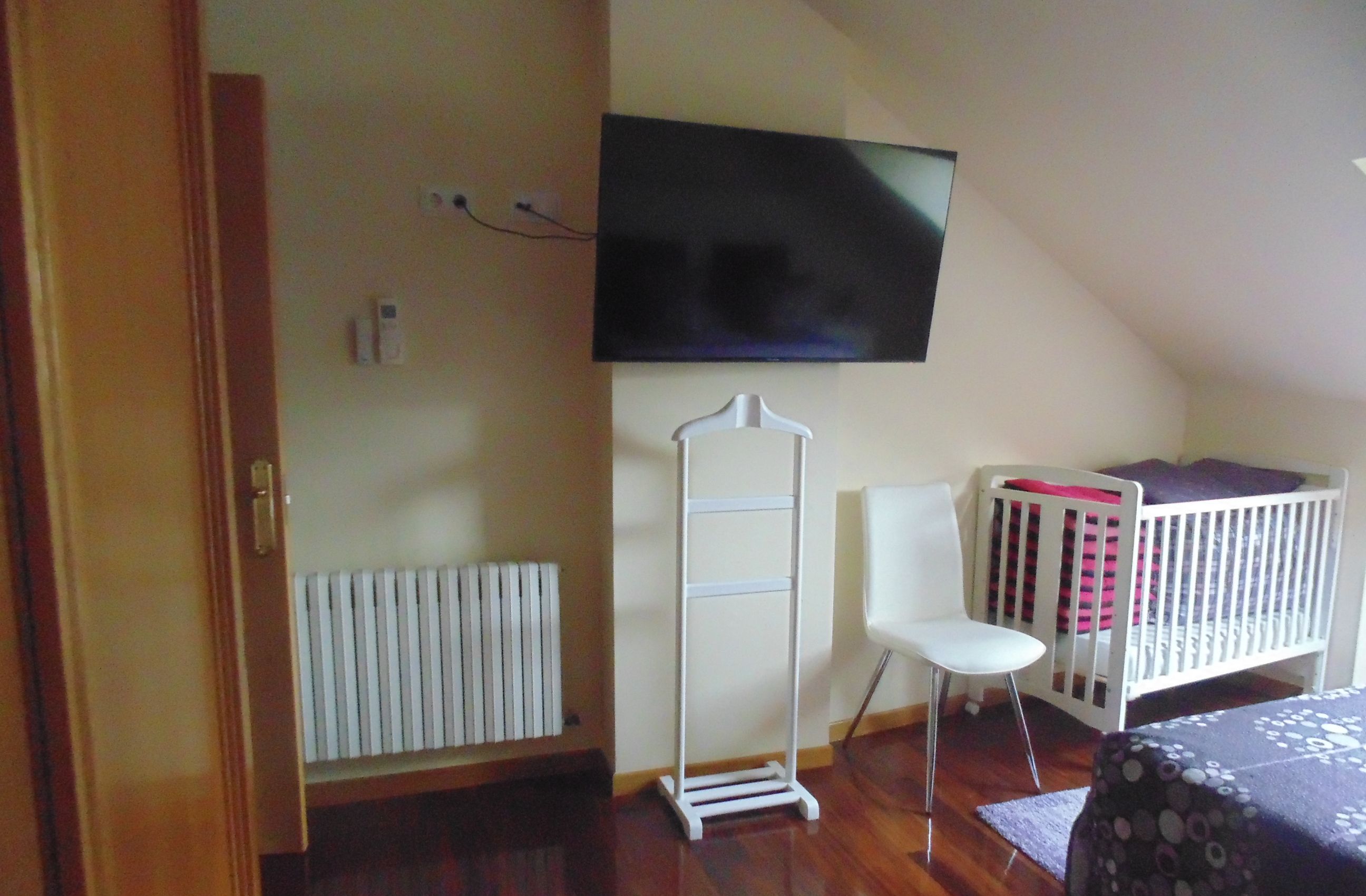 A room with a television mounted on the wall, a white radiator, a white chair, a crib, and a wooden floor.