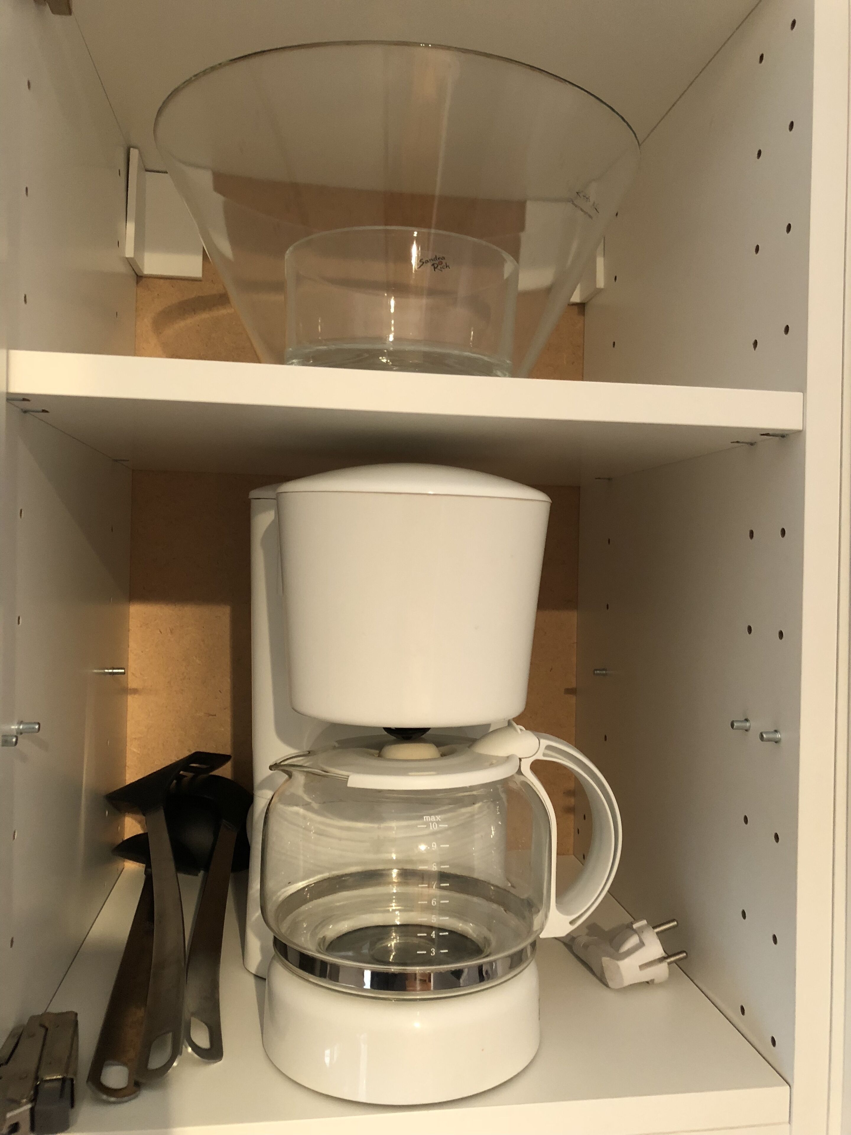 A white coffee maker with a transparent water reservoir and a cup holder on a shelf.