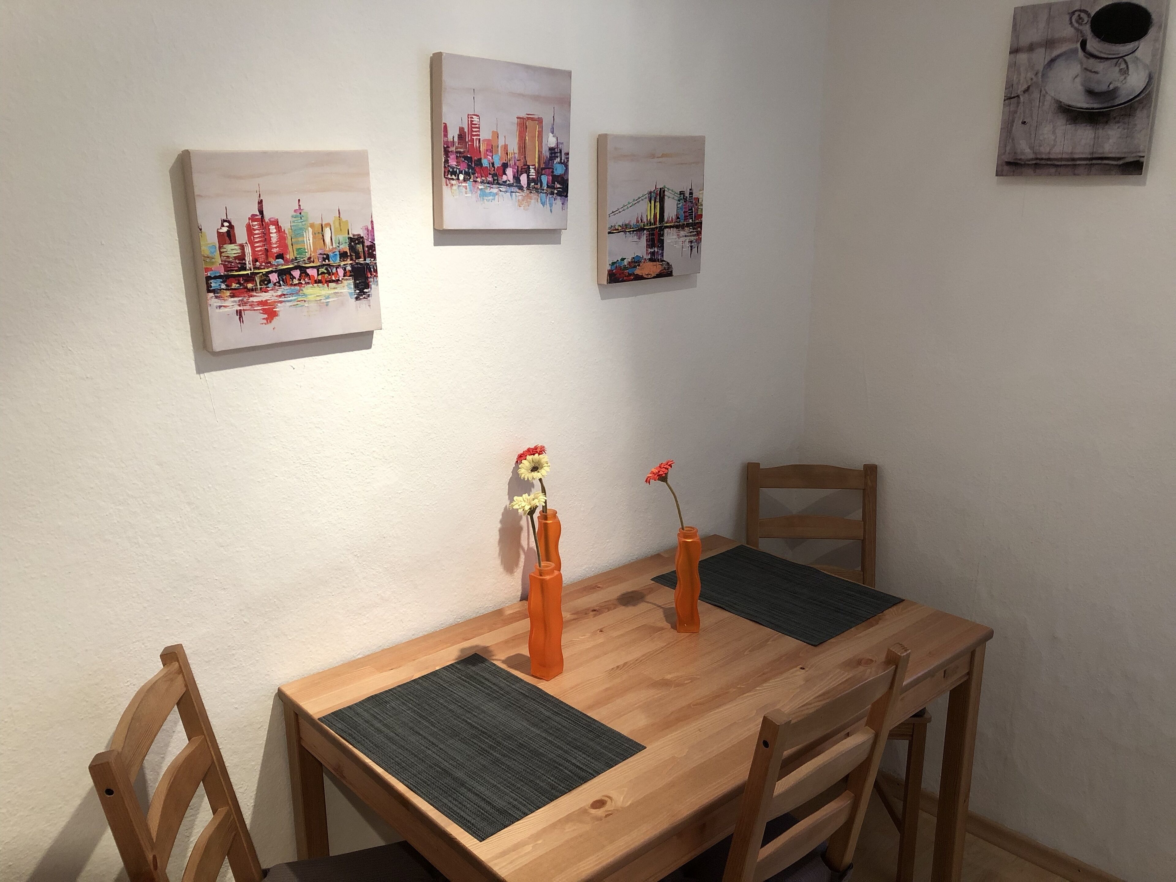 Uma mesa de jantar de madeira com duas cadeiras, um vaso com flores e três obras de arte emolduradas na parede.