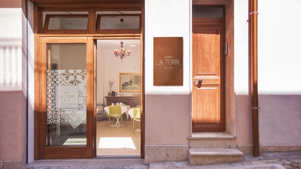 Une entrée d’immeuble avec une porte en bois et des portes vitrées, une enseigne indiquant « LA TORRE B&B », et un espace de restauration visible à l’intérieur.