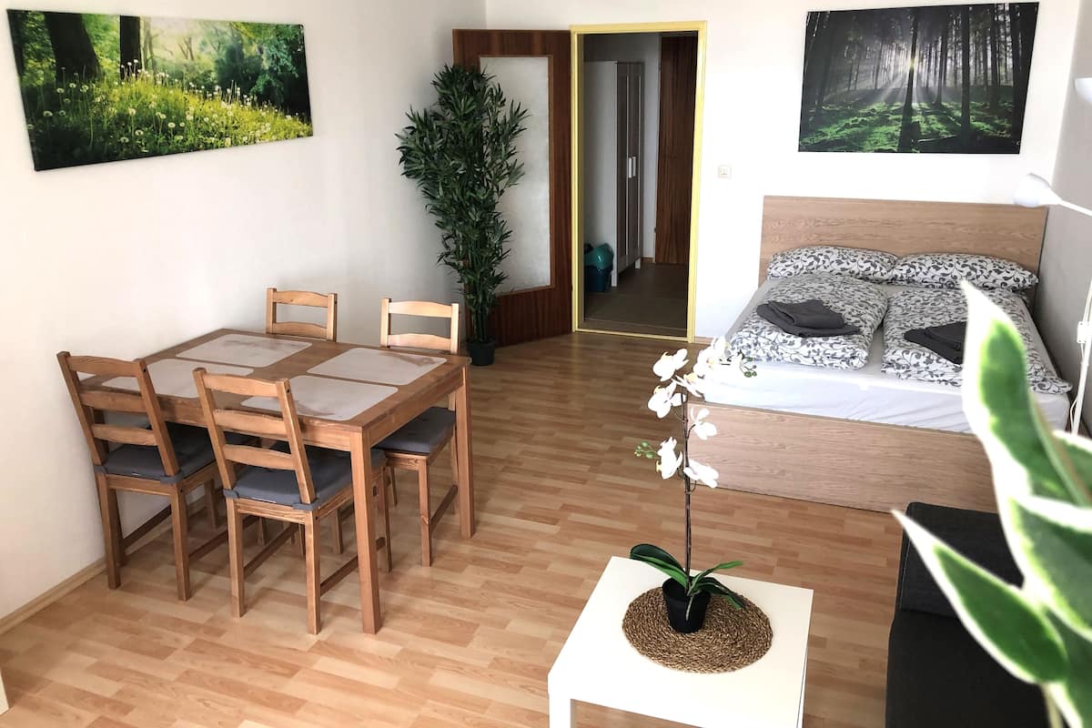 A room with a wooden dining table, chairs, a bed with patterned bedding, and two wall-mounted forest-themed paintings.