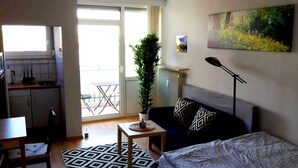 A compact living space with a kitchenette, a dining area, a sofa with patterned cushions, a small table with a decorative item, and a large window offering a view of the outdoors. - Newstyle (Regensburg)