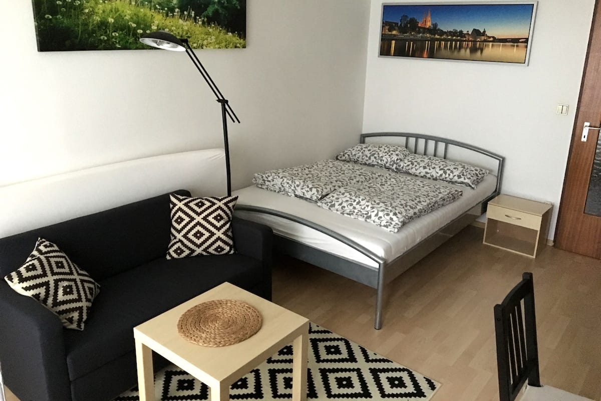 A small, modern bedroom with a bed, a black sofa, a floor lamp, and two wall-mounted pictures.