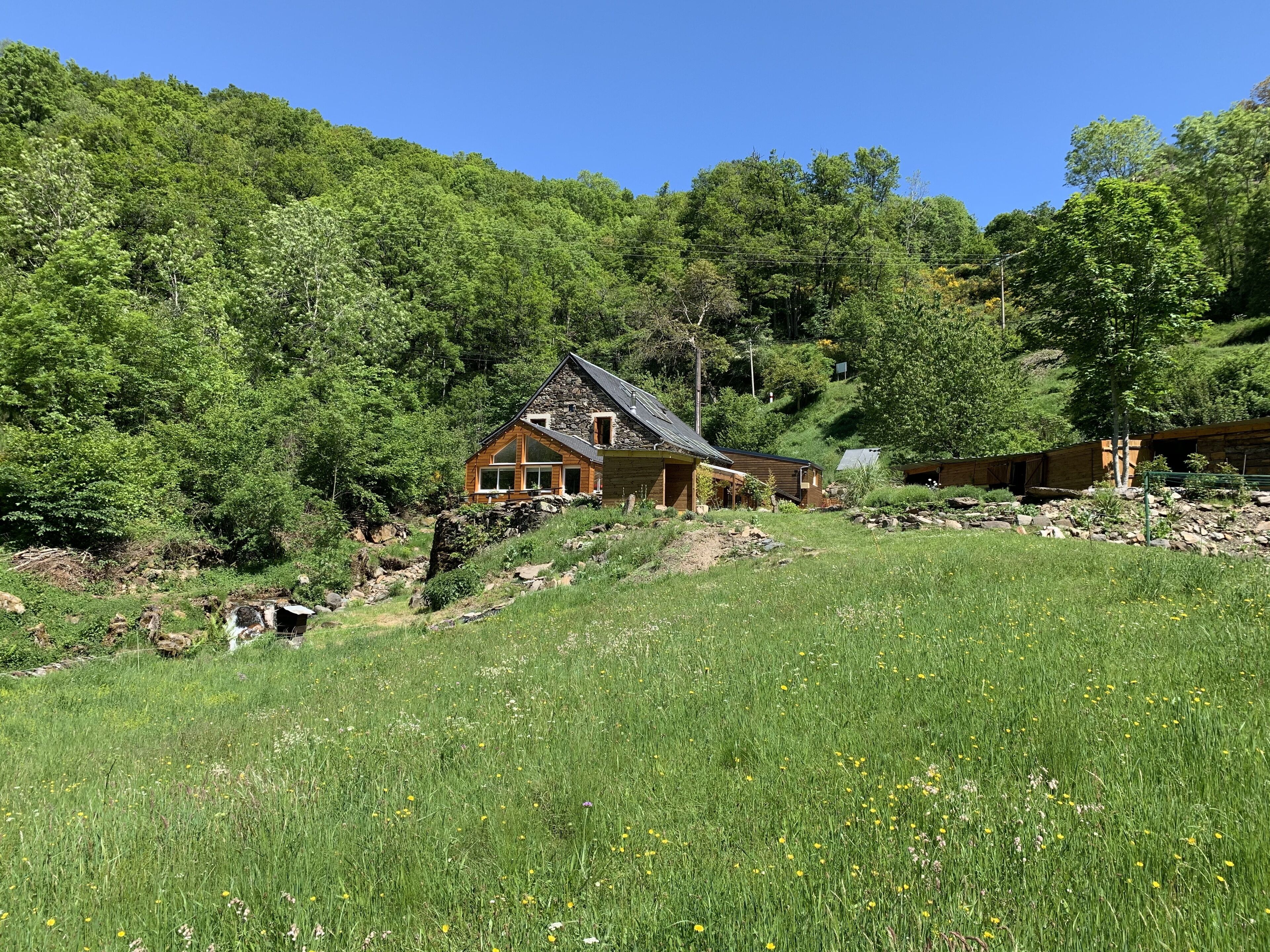 Un chalet rustique aux fondations en pierre, entouré d’une végétation luxuriante et sous un ciel bleu azur.