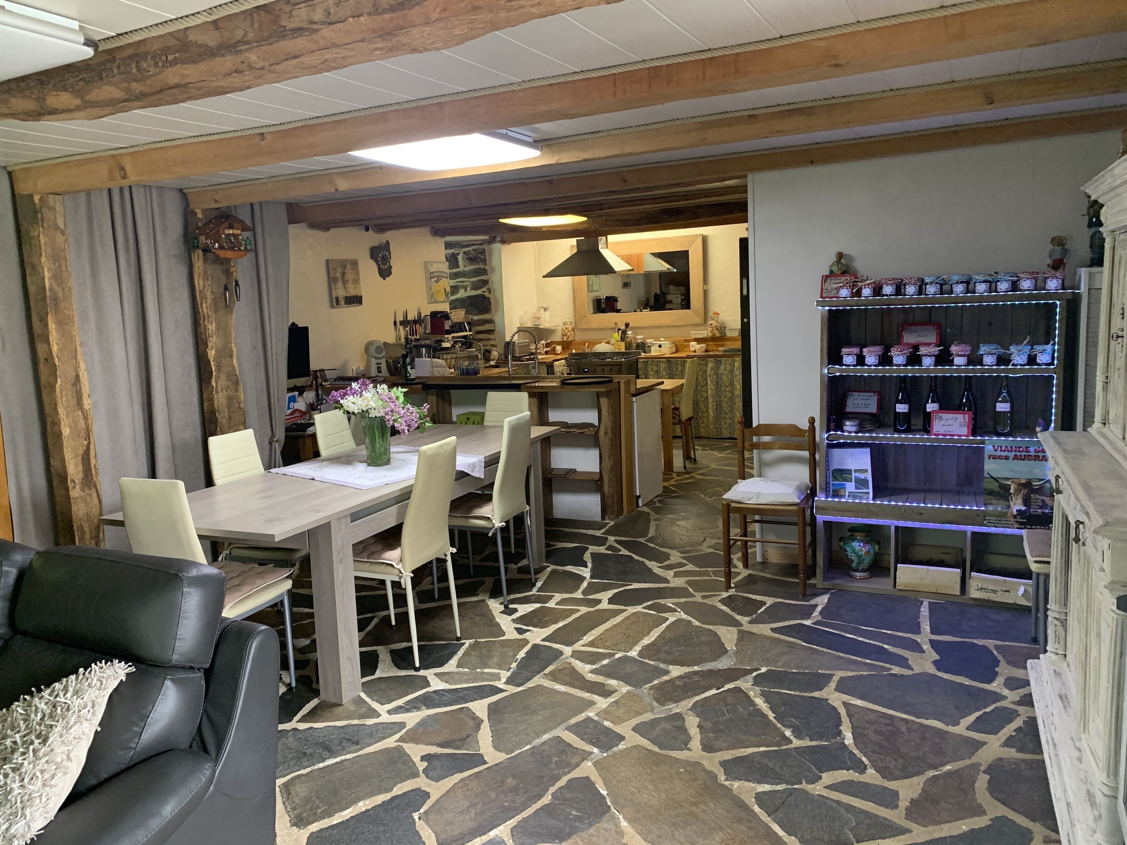 A dining area with a stone floor, a wooden table, and chairs. There is a kitchen in the background with a counter and various kitchen items.