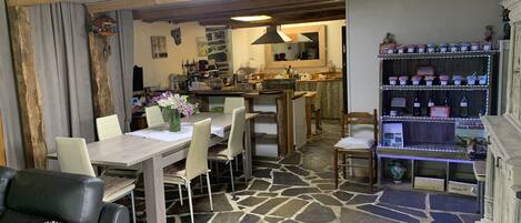 A dining area with a stone floor, a wooden table, and chairs. There is a kitchen in the background with a counter and various kitchen items.