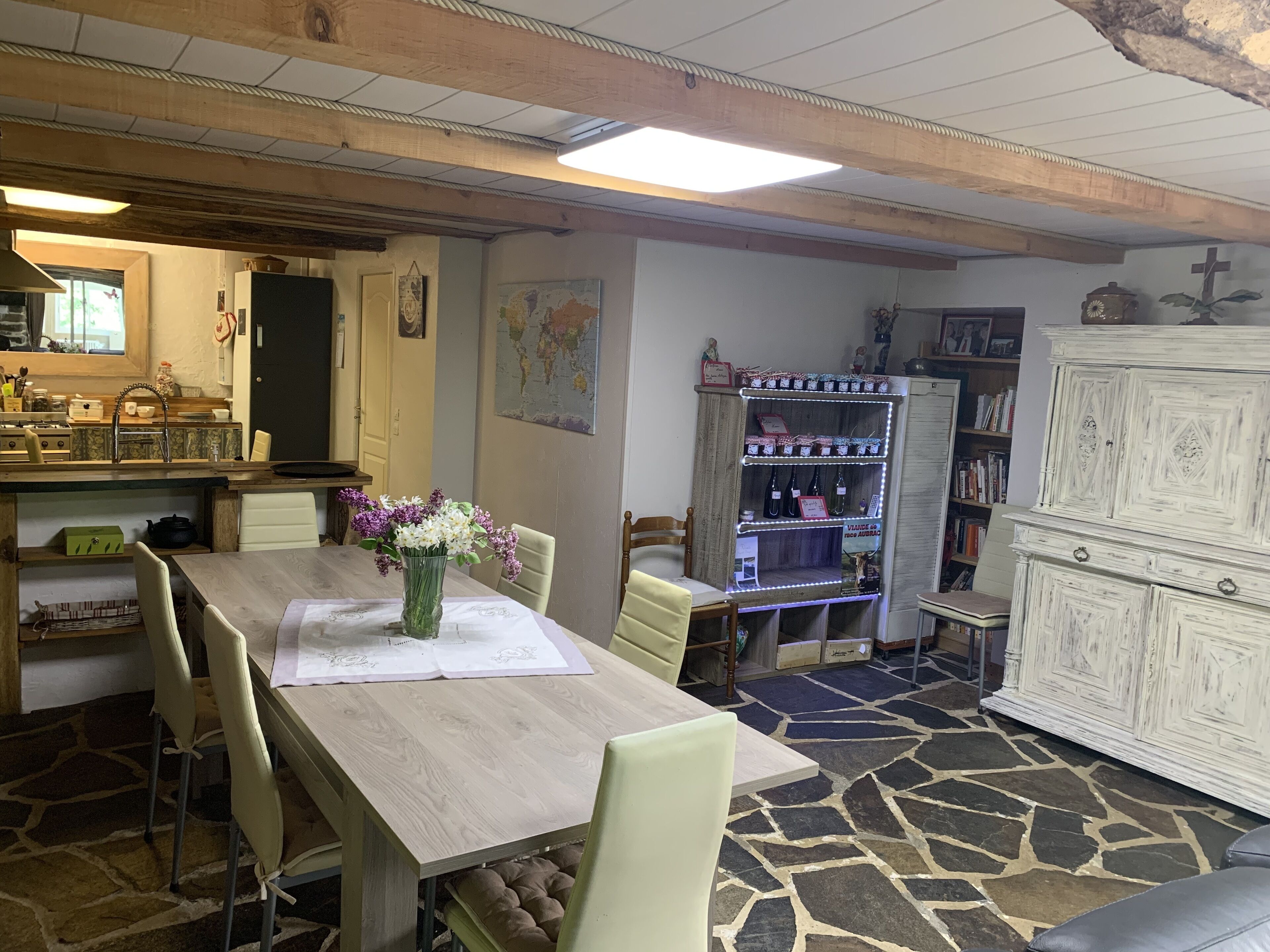 A dining area with a wooden table, chairs, a vase of flowers, a map on the wall, a shelf with bottles, and a cabinet.