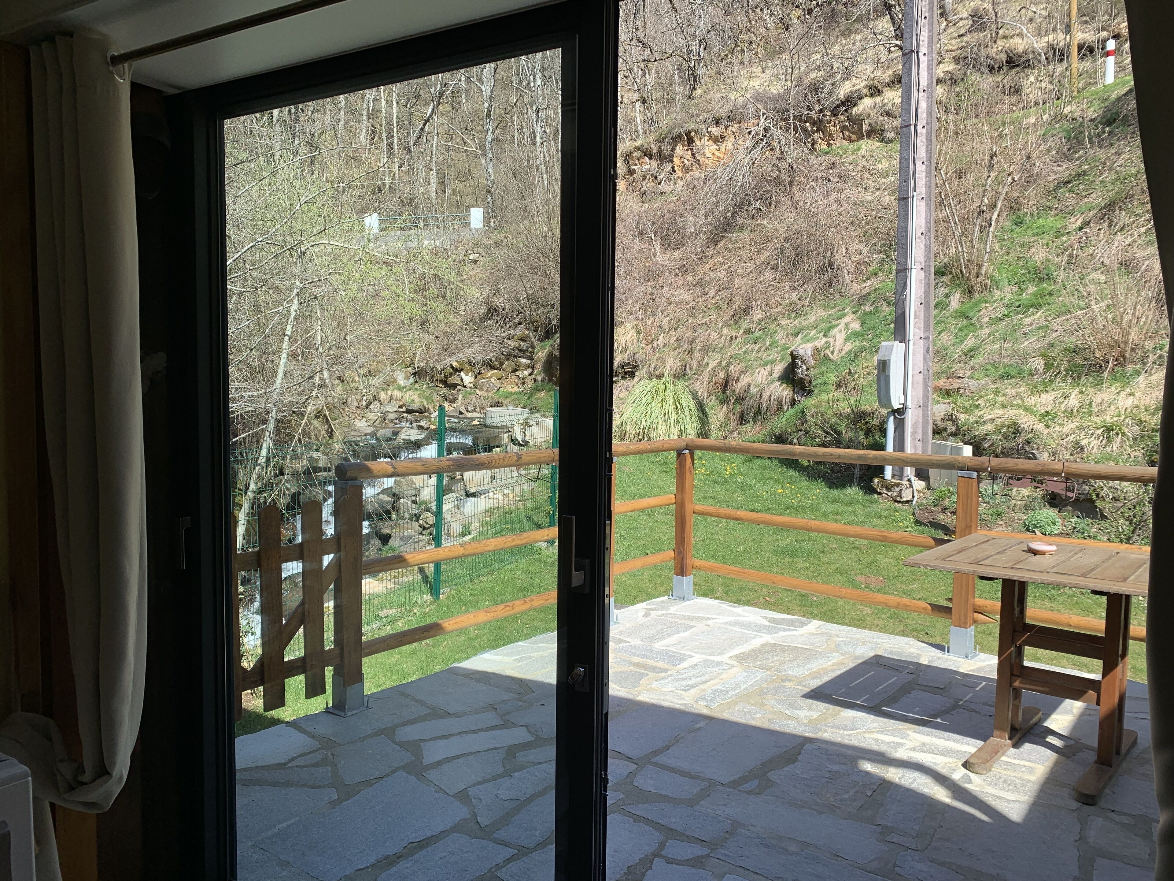 Een balkon met een stenen vloer, een houten tafel en uitzicht op een natuurlandschap met bomen en een heuvel.