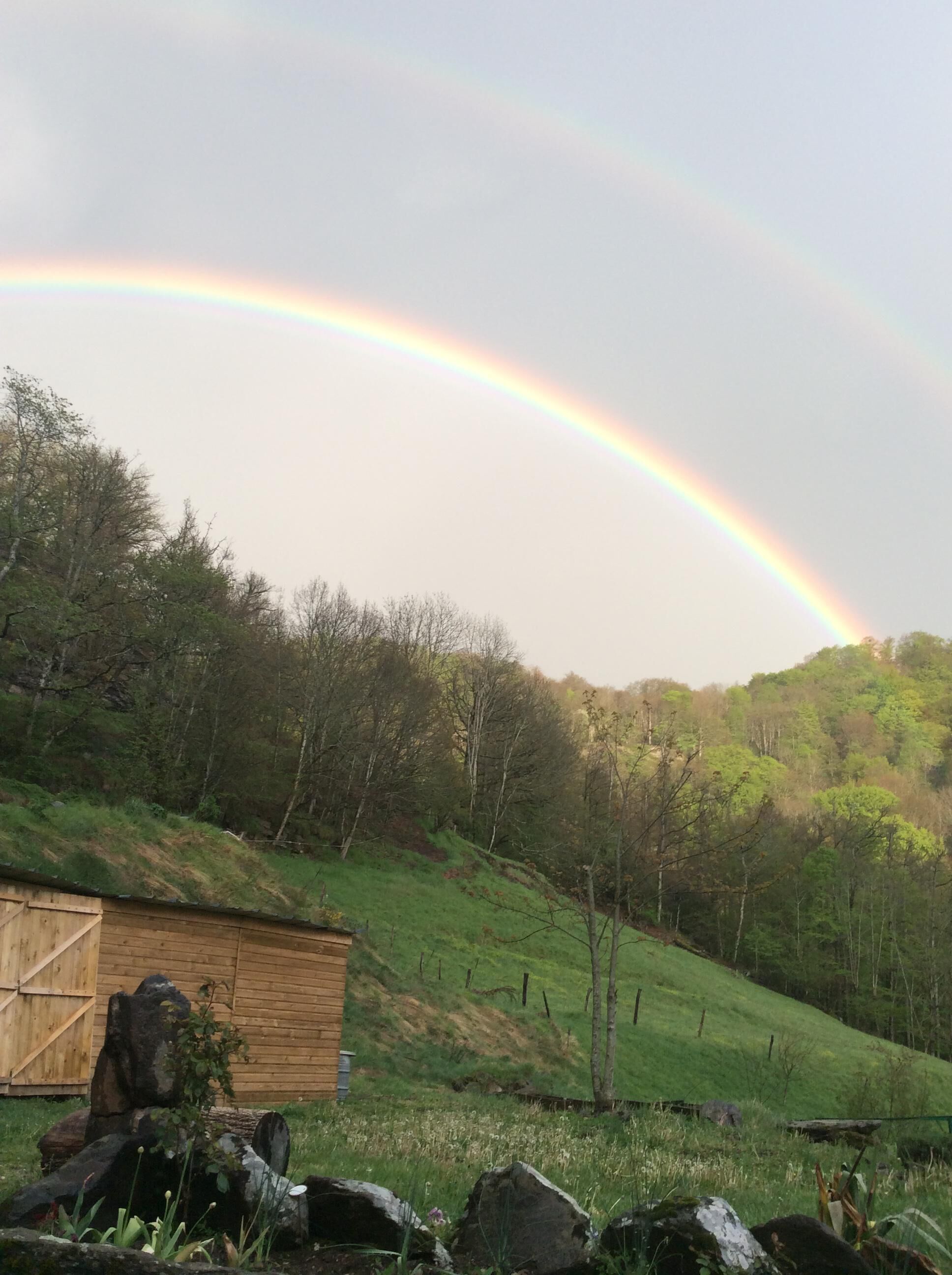 Un paysage rural avec un arc-en-ciel, une remise en bois et une colline couverte d’herbe.
