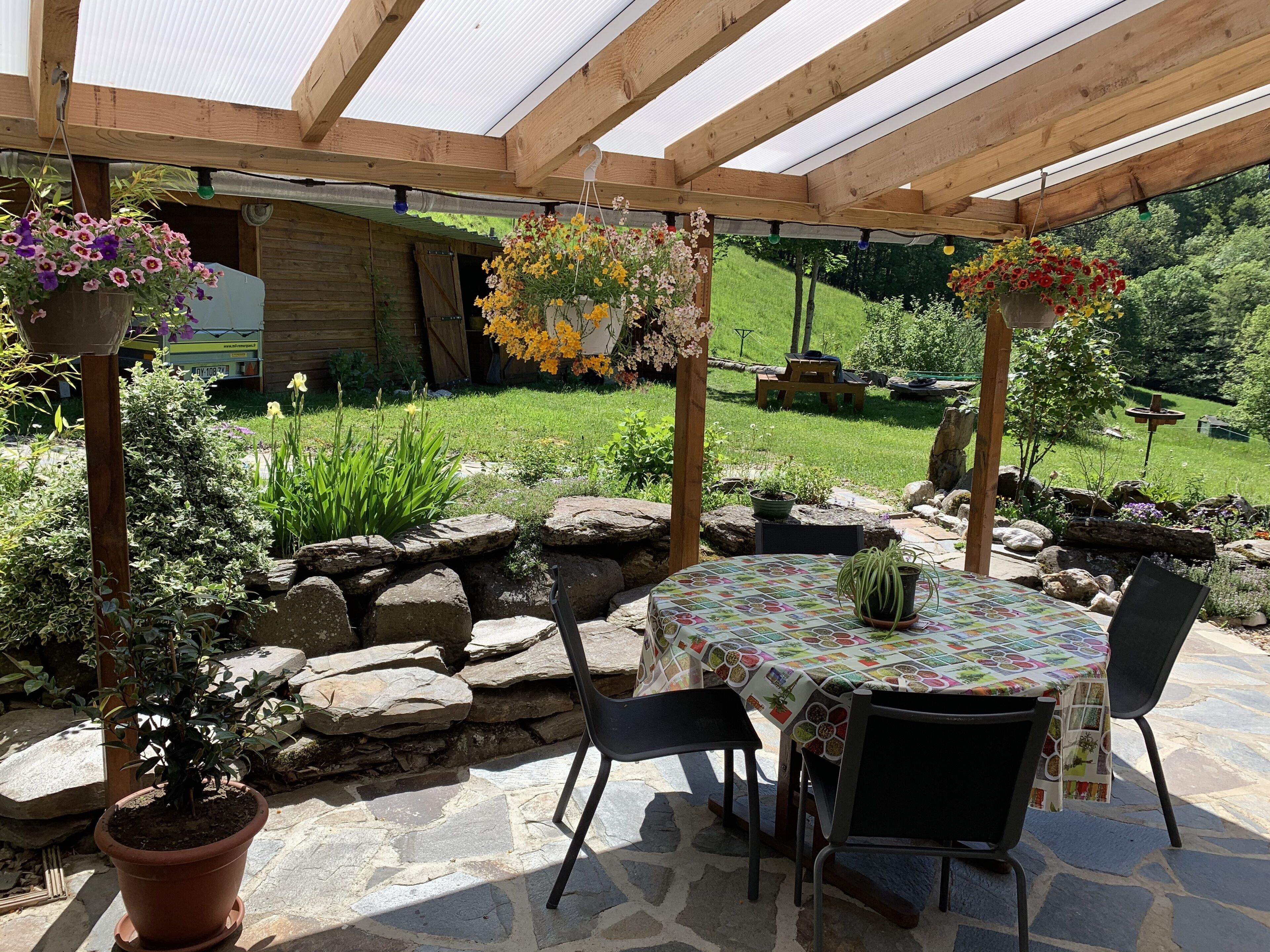 Un patio al aire libre con una mesa y sillas, rodeado de macetas con plantas y cestas colgantes de flores.